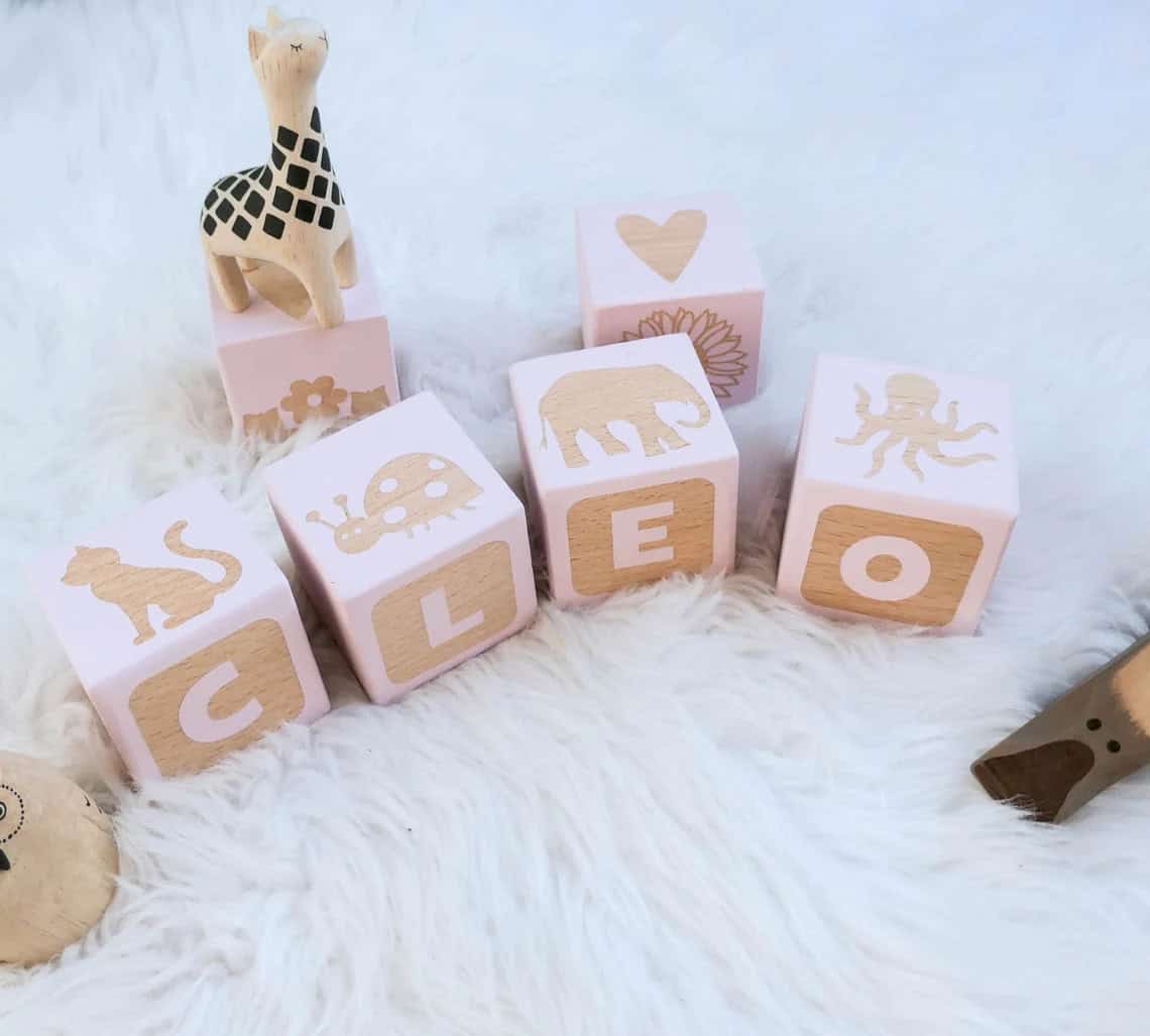 Wooden blocks on a soft white surface spell out "CLEO," with additional blocks featuring designs such as a giraffe, a heart, a flower, a ladybug, an elephant, an octopus, and a cat—an ideal DIY baby shower gift or addition to your collection of handmade baby gifts.