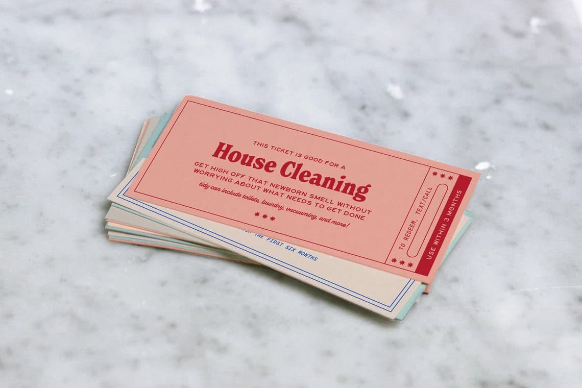 A stack of house cleaning gift certificates is displayed on a marble surface. The top certificate, pink and white with red text, offers a house cleaning service. The text is partially legible, detailing service details and terms—perfect for pairing with diy baby shower gifts.