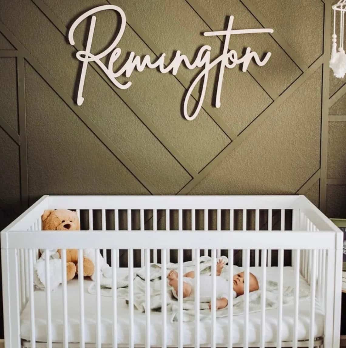 A baby sleeps in a white crib with a teddy bear and a blanket. The wall behind is decorated with a geometric pattern and features the name "Remington" in large, cursive letters. Among the cozy, modern decor, handmade baby gifts add an extra touch of warmth to the room's ambiance.