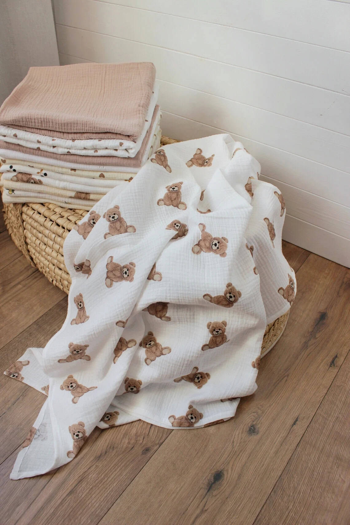 A stack of folded baby blankets in neutral tones sits on a wicker basket, showcasing perfect handmade baby gifts. One unfolded blanket with a teddy bear pattern drapes over the side of the basket and onto the wooden floor, adding charm to the room's white paneled walls.