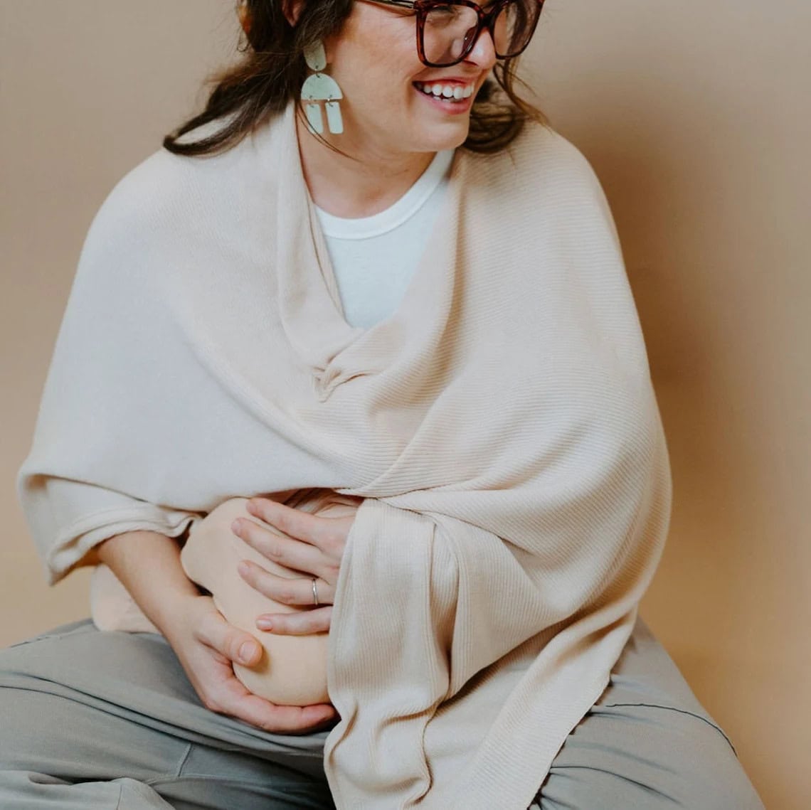 A person with glasses is seated against a beige background while breastfeeding a baby. They are wearing a white top and beige shawl, complemented by large decorative earrings. Nearby, a table displays an array of handmade baby gifts, adding to the warmth of the scene.