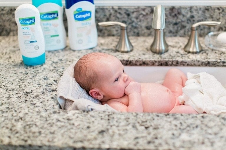A baby is lying in a granite sink, enjoying their first bath. The baby is partially wrapped in a towel and surrounded by Cetaphil Baby products. The counter is made of grey granite, with silver faucets and other scattered toiletries in the background.