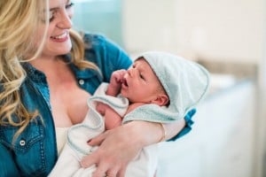 A woman with blonde hair wearing a denim shirt is holding a newborn baby wrapped in a white towel with a light blue hood covered in white polka dots. The woman, embracing her sanity as a new mom, is smiling at the baby, who appears to be looking up at her.