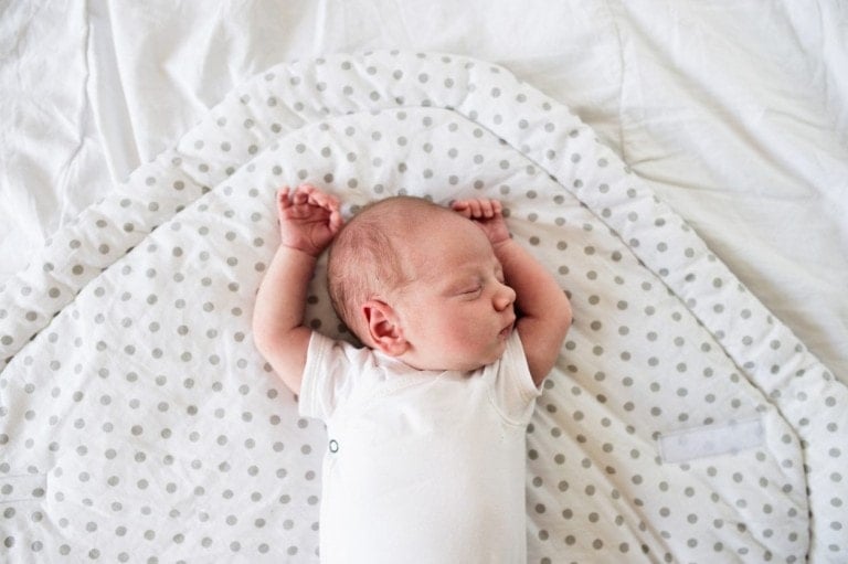 A baby with torticollis is sleeping on their back with both arms stretched upwards. The baby is lying on a white blanket with grey polka dots and wearing a white onesie. The background is a plain white sheet.