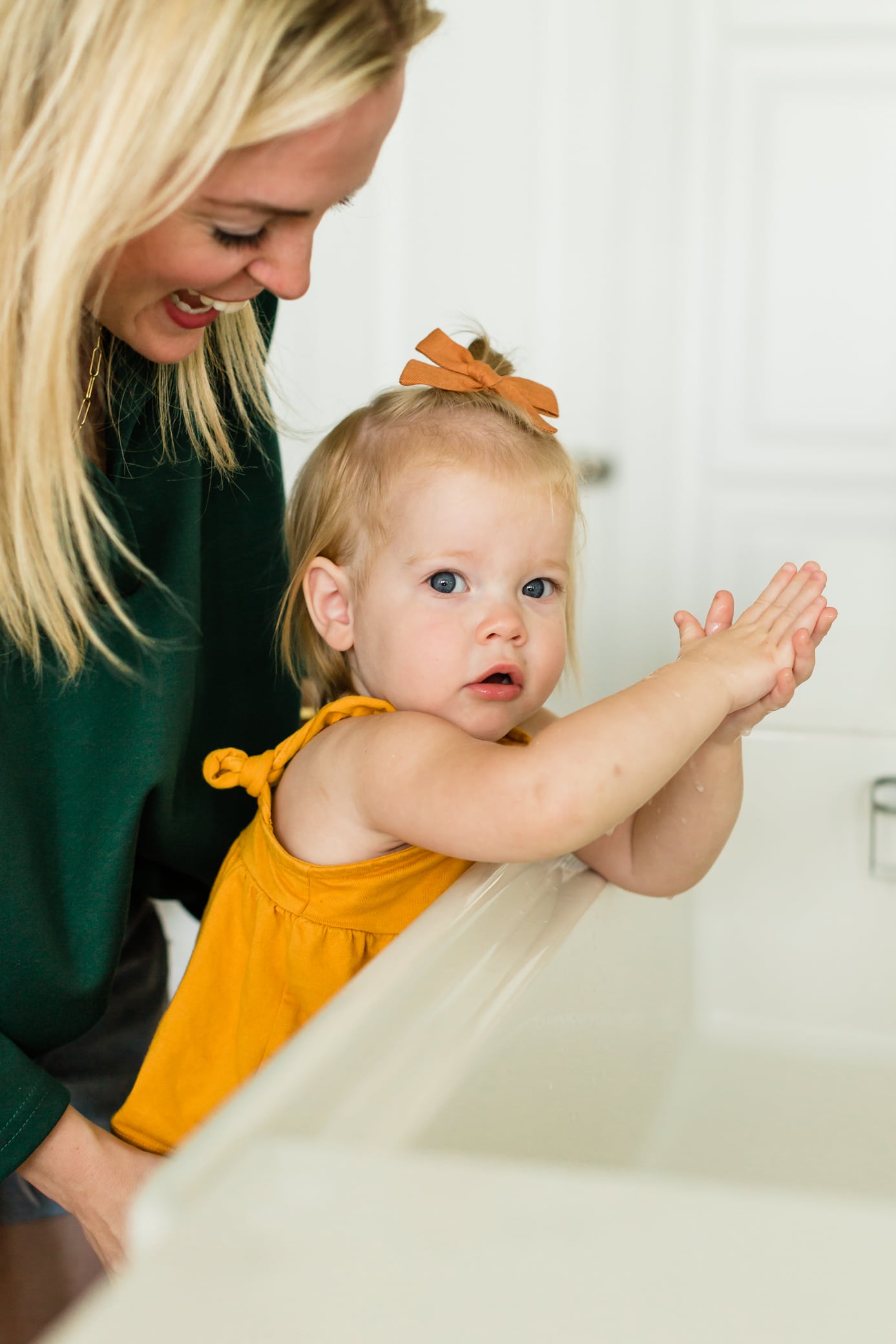Little toddler girl looking at the camera standing at the sink washing her hands with her mom behind her.
