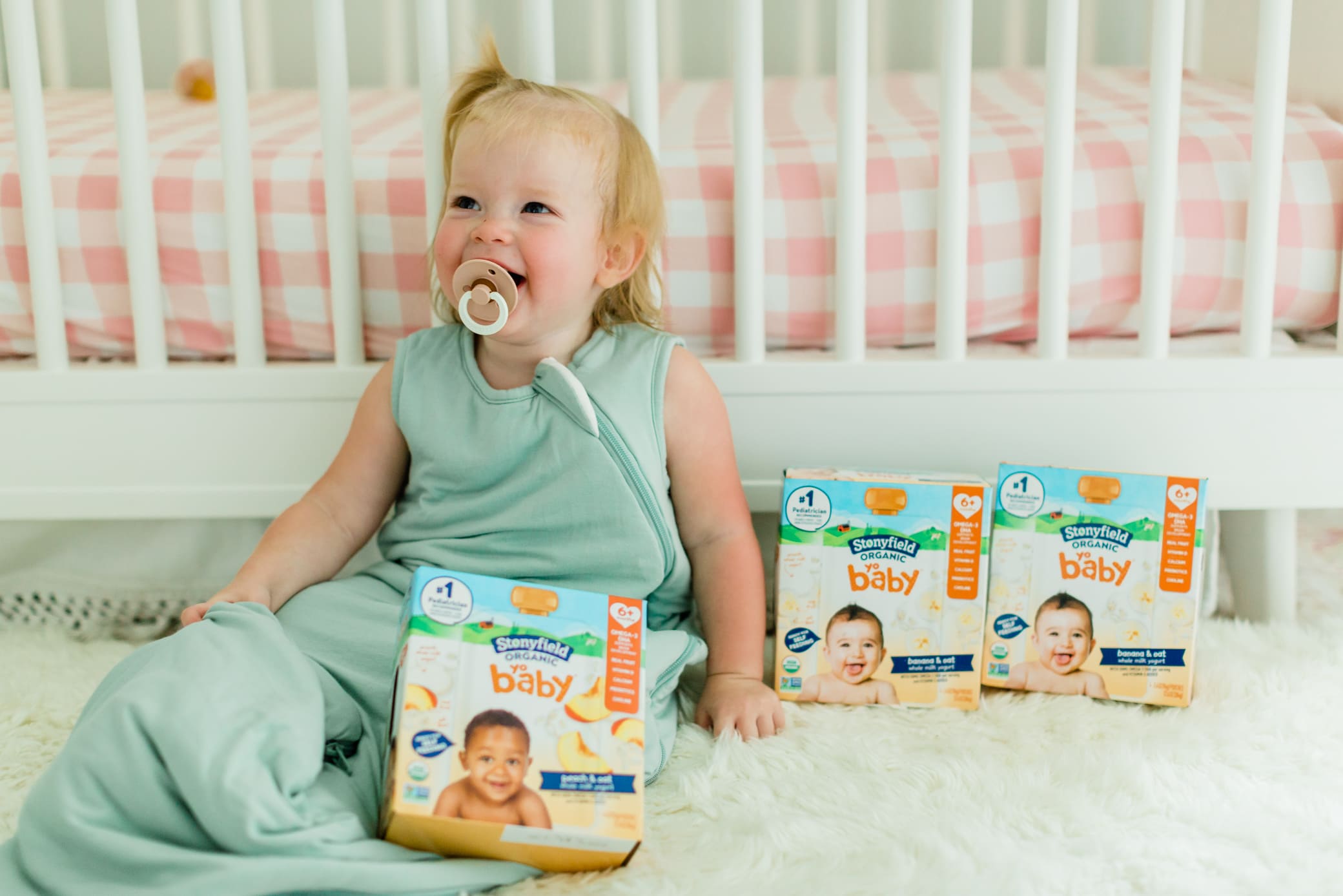 Young toddler girl sitting beside her crib wearing her sleep sack with her boxes of Stonyfield yogurt next to her.