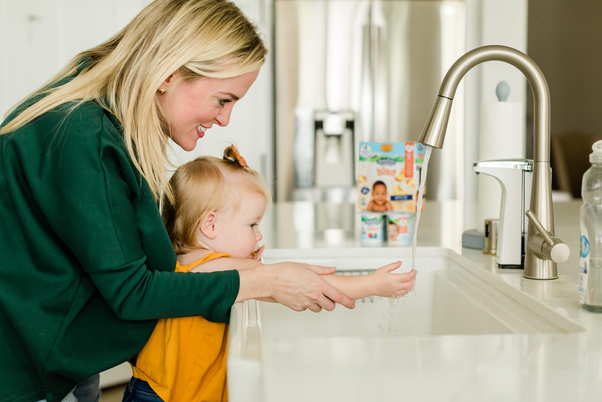 Moms standing behind her young daughter as she helps her wash her hands at the kitchen sink.