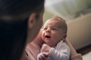 A first baby wearing a white outfit looks up while being held by an adult. The baby's mouth is open, and they appear to be happy or laughing. The adult's face is not fully visible as they look down at the baby. The background includes a radiator and some daylight.