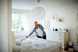 A person with short blonde hair and wearing a denim shirt over a white top is sharing quick housekeeping tips on the phone while making a bed in a bright, modern bedroom. The room features white furniture, framed photos, a decorative light fixture, and a window with a view of greenery.