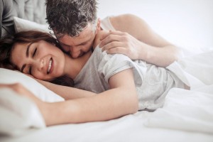 A man and a woman are lying in bed together. The man is embracing the woman from behind while smiling, and the woman is lying on her side, eyes closed with a serene smile. They appear relaxed and content, their intimate mood reflecting a renewed postpartum sex drive amidst the white bedding.
