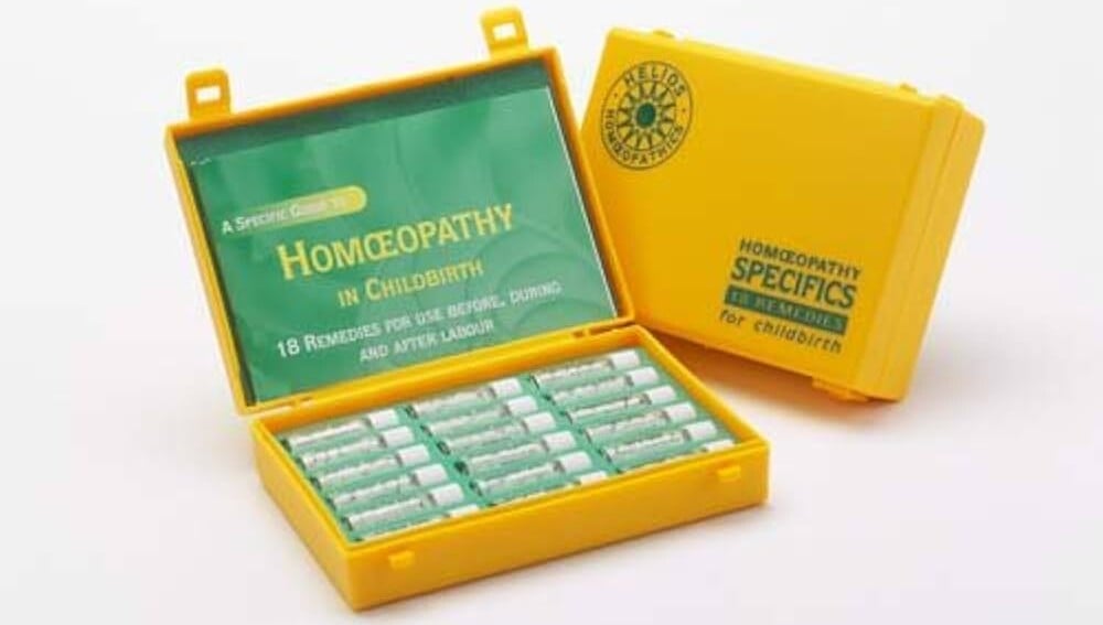 A yellow plastic case with a hinged lid is open, revealing small tubes labeled in green text. The interior lid has a green card titled Homeopathy in Childbirth, offering information on how to prepare your body for labor and birth. The exterior displays the text Homeopathy Specifics.