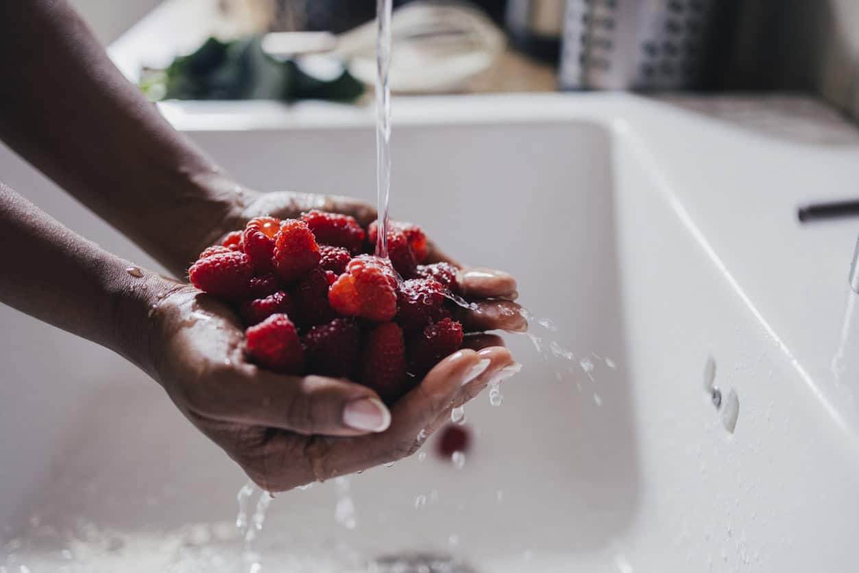 Hands holding a bunch of raspberries under running water in a kitchen sink, preparing one of the best foods to eat while pregnant—ideal for healthy pregnancy meals and nutritious snacks.