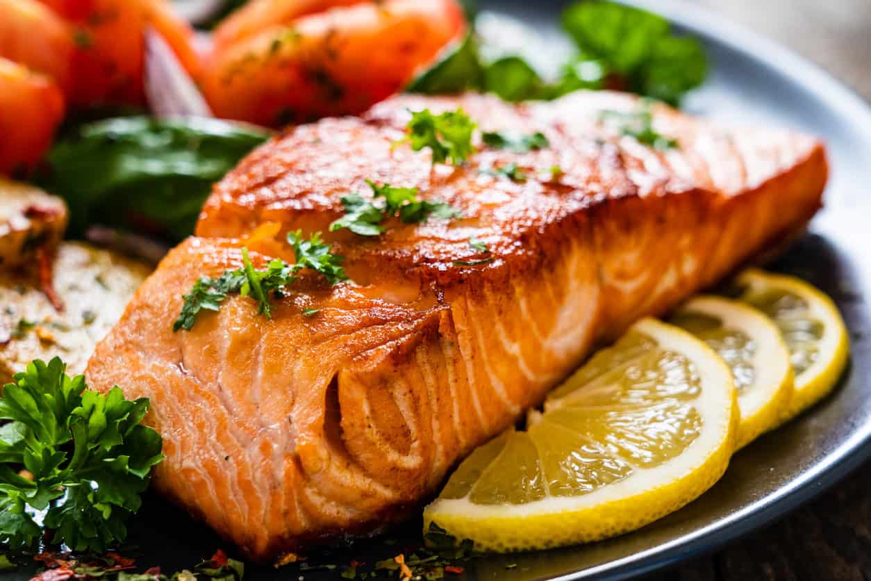 A close-up of a cooked salmon fillet, one of the best foods for pregnancy, garnished with fresh parsley. Served with slices of lemon on the side and accompanied by tomatoes and greens on a dark plate, its a delicious choice for healthy pregnancy meals.