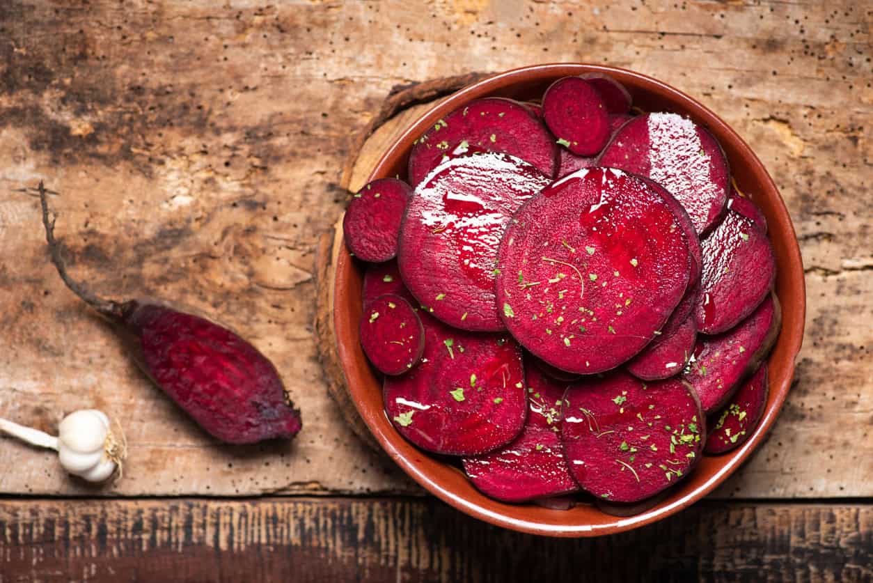 A bowl of sliced beets garnished with herbs sits on a rustic wooden surface. Beside it, a whole beet and a bulb of garlic are placed on the table, adding to the earthy setting—perfect for healthy pregnancy meals or as one of the best foods to eat while pregnant.