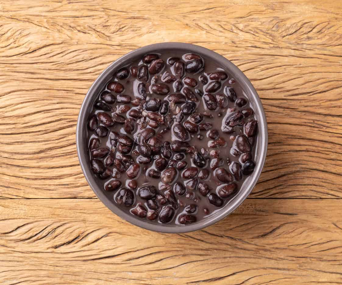 A bowl filled with black beans in liquid rests on a textured wooden surface. As one of the best foods for a healthy pregnancy, the beans, evenly covered with liquid, sit atop the natural, wavy grain pattern of the wood background.