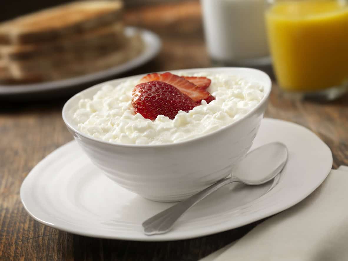 A bowl of cottage cheese topped with a sliced strawberry sits on a white plate with a silver spoon. In the background, theres a stack of toast, a glass of milk, and a glass of orange juice on a wooden table—ideal elements for healthy pregnancy meals.