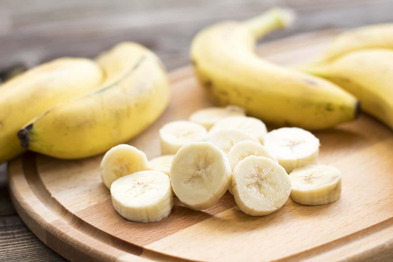 A wooden cutting board showcases sliced bananas in the foreground, perfect for healthy pregnancy meals, with whole bananas in the background. The light brown surface and kitchen setting create a warm atmosphere, ideal for preparing the best foods to eat while pregnant.