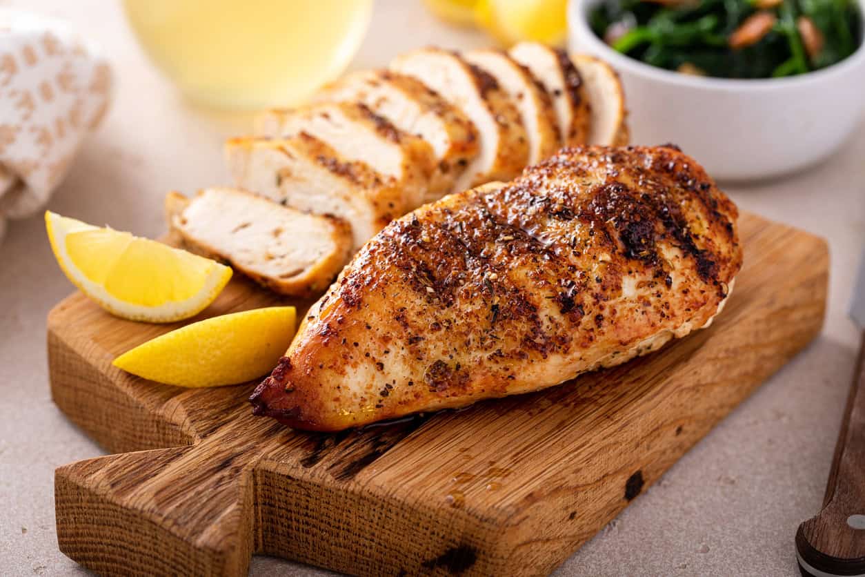 Grilled chicken breast seasoned with spices rests on a wooden cutting board, accompanied by lemon wedges—perfect for the best foods to eat while pregnant. Behind are chicken slices and a bowl of greens. A glass of white wine is partially visible in the background.