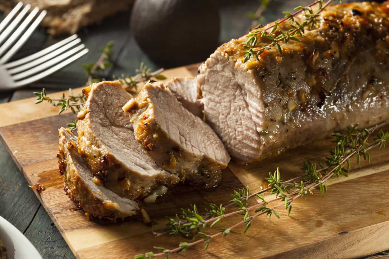 A cooked pork loin is shown on a wooden cutting board. Partially sliced with several pieces visible, this nutritious option is perfect for meals—especially when considering the best foods to eat while pregnant. The meat is garnished with fresh thyme, and there are forks placed in the background.
