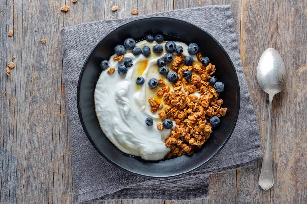 A black bowl holds yogurt topped with granola, fresh blueberries, and a drizzle of honey—a perfect healthy snack for pregnancy. Resting on a gray cloth napkin on a wooden table, the scene is completed with a silver spoon placed to the right.