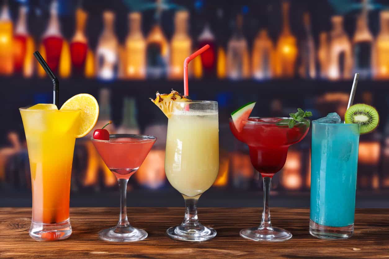 Five colorful cocktails are arranged on a wooden bar counter, each in a distinct glass with garnishes like orange slices, cherries, pineapple, and kiwi. Blurred bottles are visible in the background.