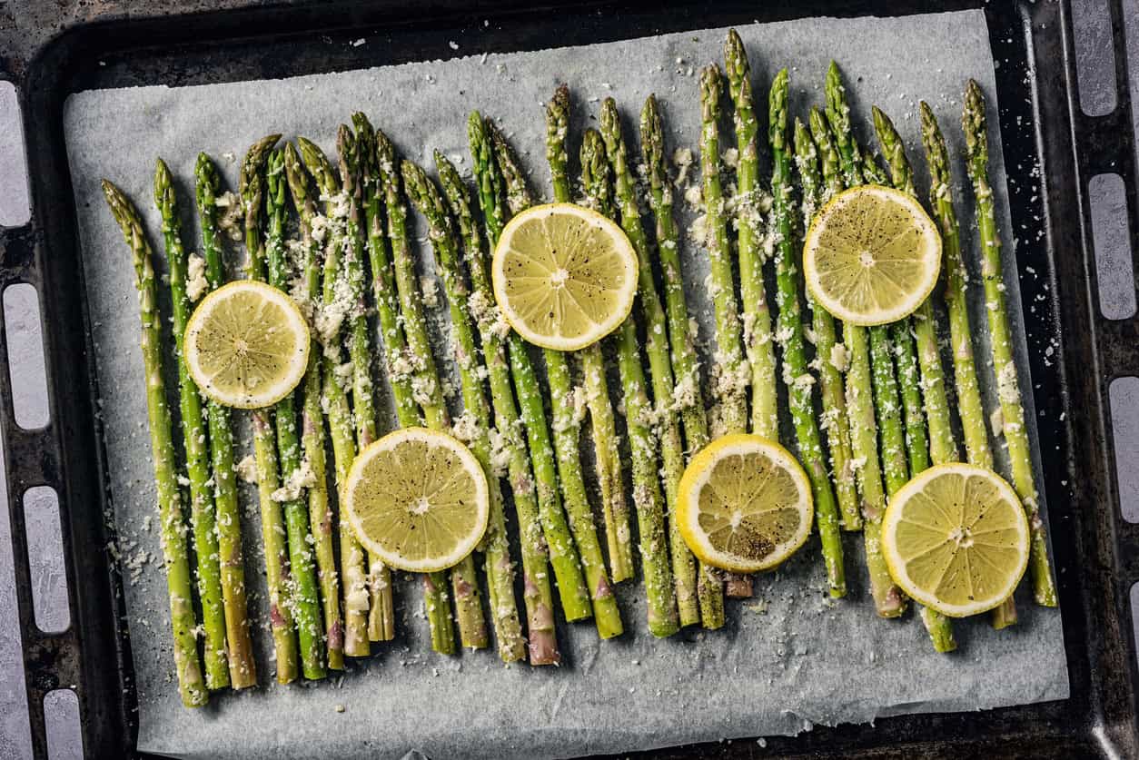 A baking tray lined with parchment paper holds fresh asparagus spears, one of the best foods to eat while pregnant, topped with grated cheese and lemon slices, ready for roasting.