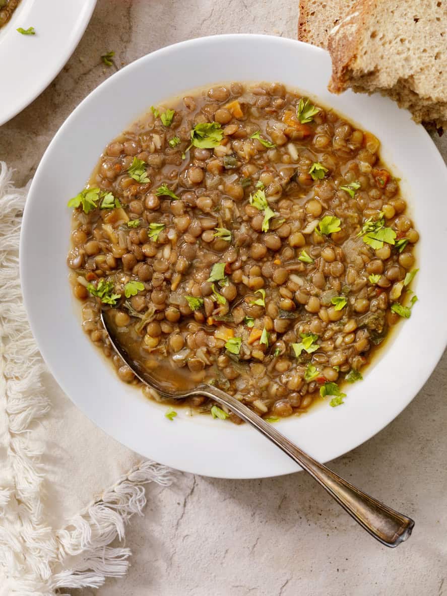 A white bowl filled with lentil soup, a nutritious choice among the best foods for pregnancy, is garnished with chopped parsley. A metal spoon rests inside the bowl. Two slices of brown bread are partially visible on the side, all set on a light-colored cloth.