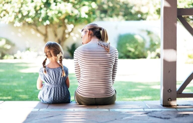 A woman and a strong-willed child with braided hair sit on a porch step, facing away from the camera. They are looking out onto a sunlit garden with grass and trees. The woman wears a striped shirt, and the child wears a blue dress with polka dots.