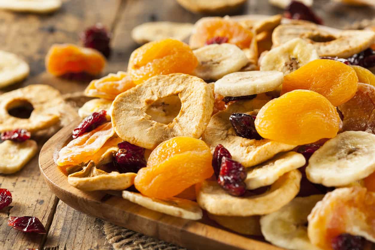 A wooden plate filled with assorted dried fruits, including apricots, apple rings, banana chips, and cranberries—ideal as part of healthy pregnancy meals—is placed on a rustic surface. Scattered fruits surround the plate.
