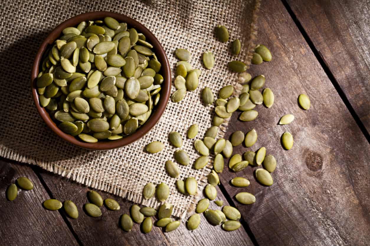 A bowl of pumpkin seeds, celebrated as one of the best foods for pregnancy, rests on a piece of burlap atop a wooden surface. A few seeds are scattered outside the bowl, enhancing the rustic presentation of this healthy snack.