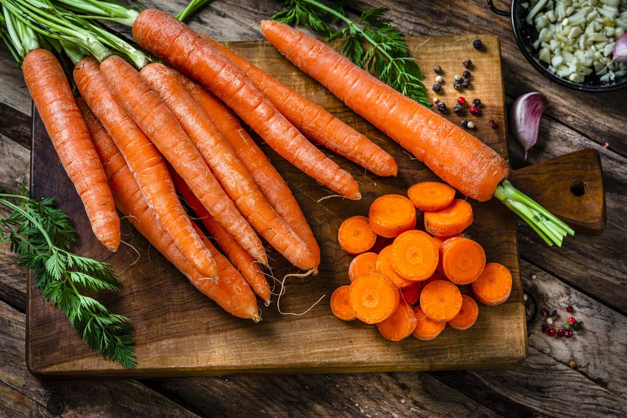 A wooden cutting board holds whole carrots and sliced rounds, with fresh greens attached. Scattered black peppercorns and garlic cloves hint at the best foods for pregnancy, as nutrient-rich veggies play a vital role in a wholesome diet while expecting.
