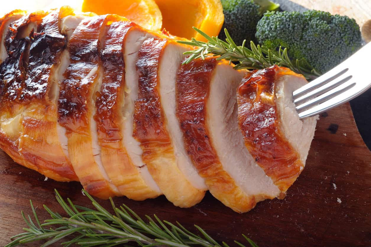 Sliced roasted turkey breast on a wooden cutting board, garnished with rosemary sprigs. A fork is inserted into one of the slices. In the background, alongside healthy snacks for pregnancy like broccoli and sliced oranges, this meal offers a nutritious option for expectant mothers.