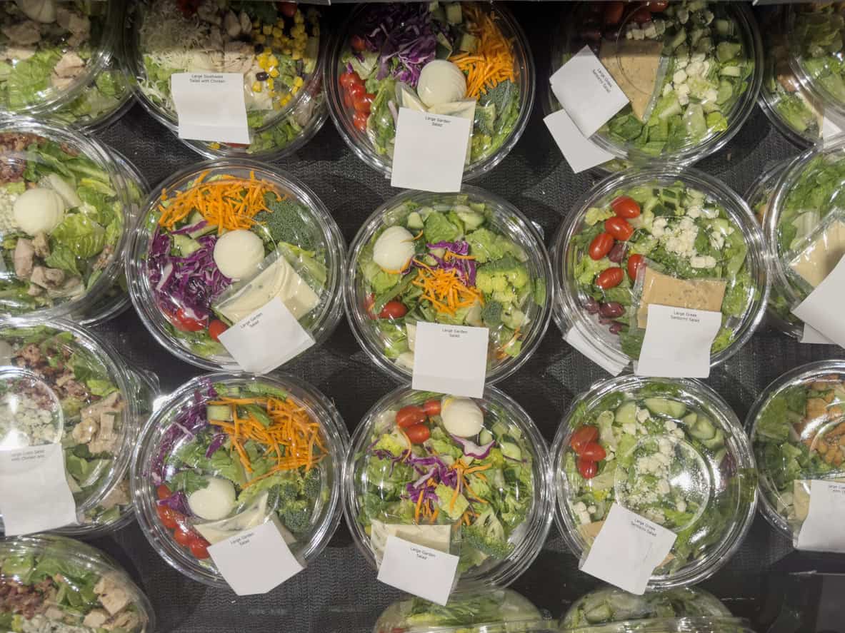A display of packaged salads in clear plastic containers, each brimming with ingredients like lettuce, cherry tomatoes, shredded carrots, hard-boiled eggs, and cheese. Perfect as healthy snacks for pregnancy, these are conveniently labeled on top for easy selection.
