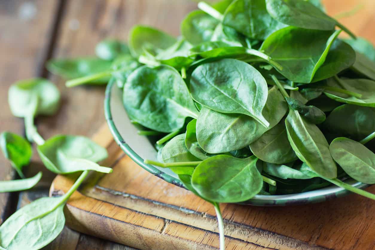 A bowl filled with fresh spinach leaves, one of the best foods to eat while pregnant, is placed on a wooden cutting board. A few spinach leaves are scattered on the surface around the bowl.