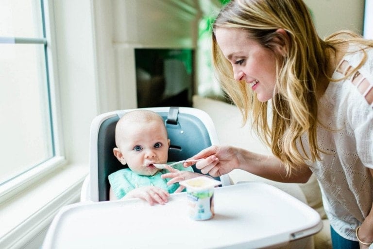 A woman is feeding her 6-month-old baby, who is seated in a high chair by the window. The baby, wearing a green bib, is being spoon-fed from a container of baby food. The woman smiles warmly as she holds the spoon, while the baby looks ahead with curious eyes.