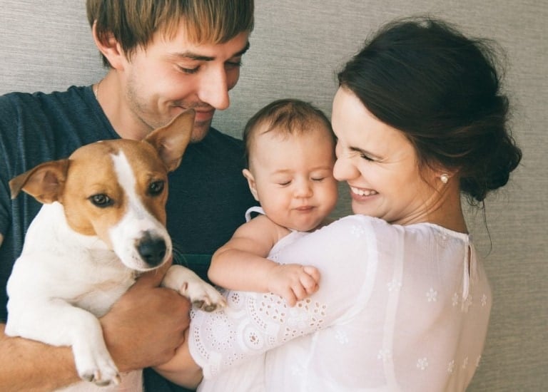 A man and woman are smiling at a baby they are holding. The man is also holding a brown and white dog, adding to the sense of hygge. The baby has its eyes closed. The group is standing against a plain background.