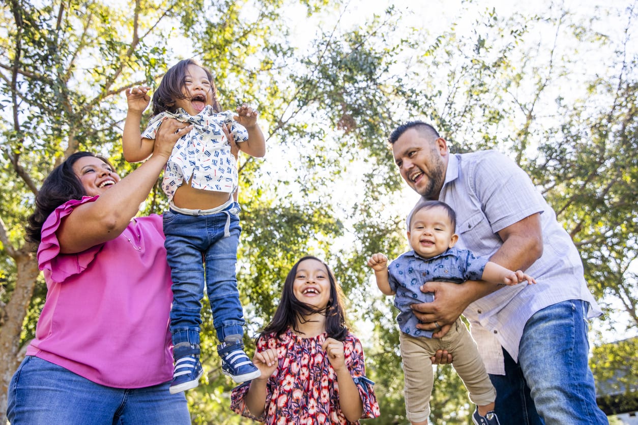 A young mexican family plays in the park, the parents lifting their smallest children up. The young boy has down's syndrome.