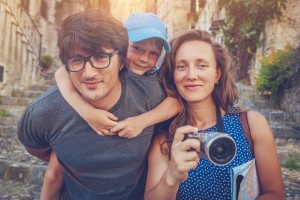 A man with glasses, a child in a blue cap on his back, and a woman holding a camera pose for DIY professional-looking family photos on a stone pathway in a historic-looking area. The background shows old buildings and greenery, suggesting they are in a picturesque village or town.
