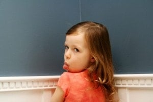 A young child with long, light brown hair is facing away from the camera, turning her head to look back over her shoulder. She is sticking her tongue out and wearing a red-orange shirt. The background consists of a blue wall with white trim, capturing a playful moment in the journey to raise a child.