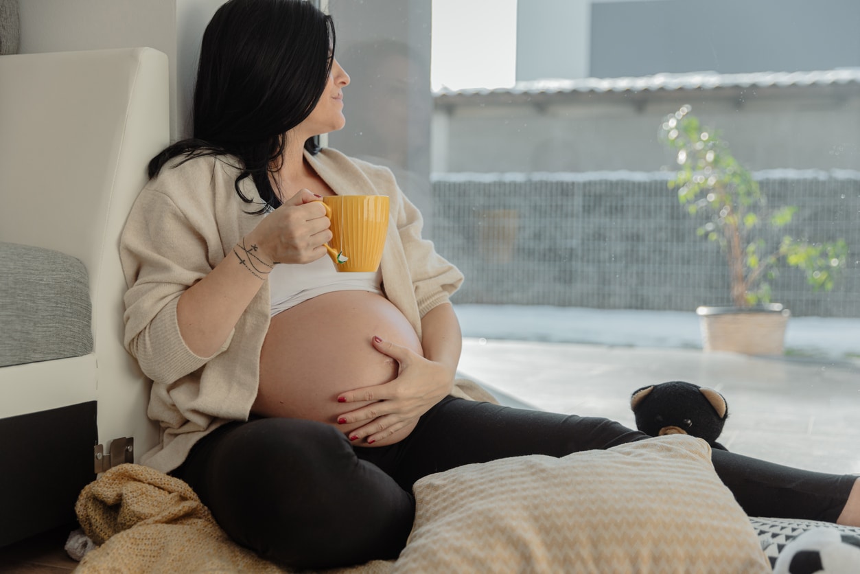 Pregnant woman drinking coffee while relaxing at home and looking out through window