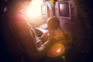 A mother and her baby girl sit in a passenger airline seat, the sun shining brightly in through the plane window. While air travel with children can be difficult, both mom and child are content, the mother with a smile on her face. Horizontal image.