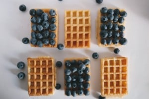 Six rectangular waffles are arranged on a white surface, with five topped with blueberries and a few loose blueberries scattered around.
