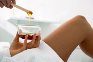A person in a white robe holds a glass bowl of wax while another hand uses a spatula to apply the wax, suggesting a waxing procedure.