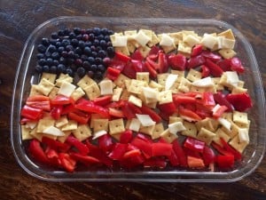 A rectangular dish contains food arranged to resemble the American flag, perfect for a Fourth of July celebration. Blueberries represent the blue field with stars, while red bell peppers and white cheese cubes are alternated in rows to symbolize the red and white stripes of the flag.