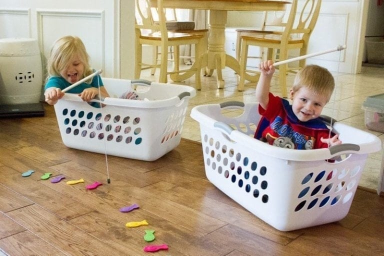 Two children are sitting inside white laundry baskets on a wooden floor, each holding a stick with a string to play a fishing game—a perfect example of indoor activities for toddlers. Colorful toy fish are scattered in front of the baskets, with a kitchen table and chairs in the background.