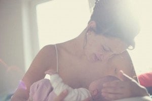 A woman is breastfeeding a baby in a softly lit room. The soothing sunlight streams in from behind, creating a warm and tranquil atmosphere. With the guidance of lactation consultants, she looks down at the baby with a gentle, caring expression.