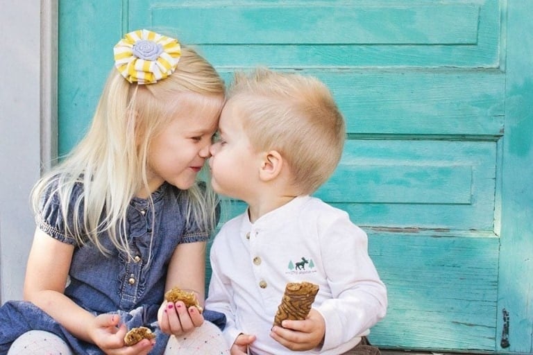 Two young children sit next to each other in front of a turquoise door. The girl on the left, wearing a denim dress and a flower hair clip, smiles warmly at the boy on the right in his white shirt. They hold pinecones and lean in close to each other, as if done having babies is still far from their minds.