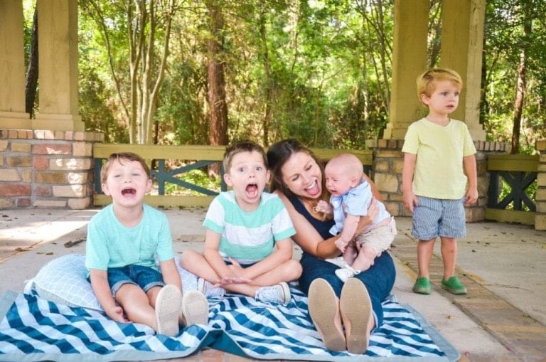 A woman and four children from a big family are sitting on picnic blankets in a gazebo. The woman holds a baby, while two boys sit next to her smiling and a third boy stands nearby, looking away. Trees and foliage are visible in the background.