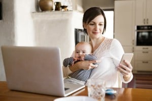 A woman is sitting at a table holding a baby with one arm while skillfully maintaining smart phone balance in her other hand. An open laptop rests in front of her beside a glass of water. The background features kitchen cabinets and various household items.