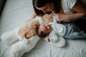 A woman lies on a bed gazing at a baby who is lying beside her on a large stuffed animal. The baby, in a white onesie and striped pants, looks up at the woman with innocent eyes. Both are on a white bedspread, creating a tranquil scene that dispels any notion of the worst advice given.